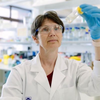 A woman stands in a laboratory wearing a white coat and glasses and holds up a glass vial